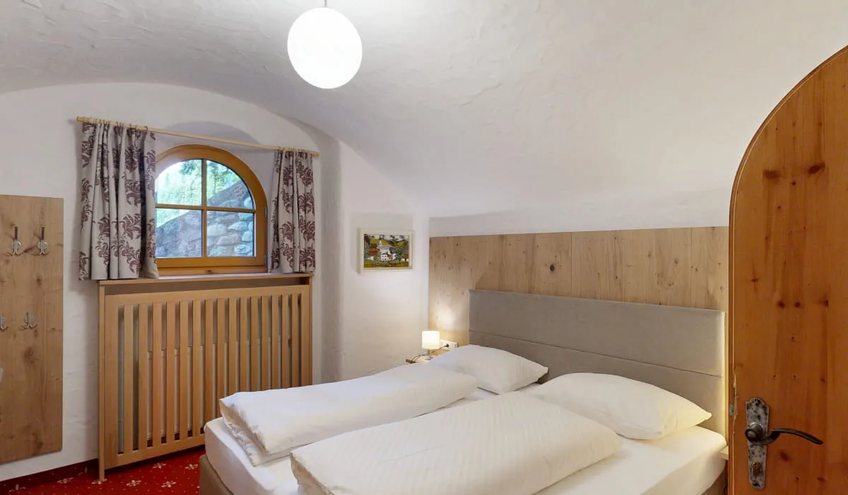 Helles Zimmer mit Doppelbett, Fenster mit Vorhängen und Holztür.